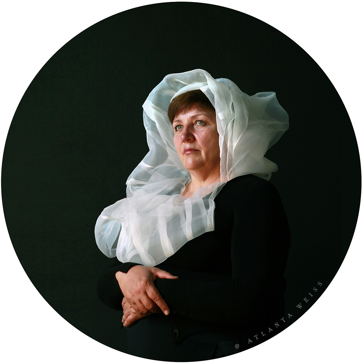 Portrait of a woman (Lucy) seated against a dark background, wrapped in a translucent white veil, circular crop, soft side light — photographic work by Atlanta Weiss.