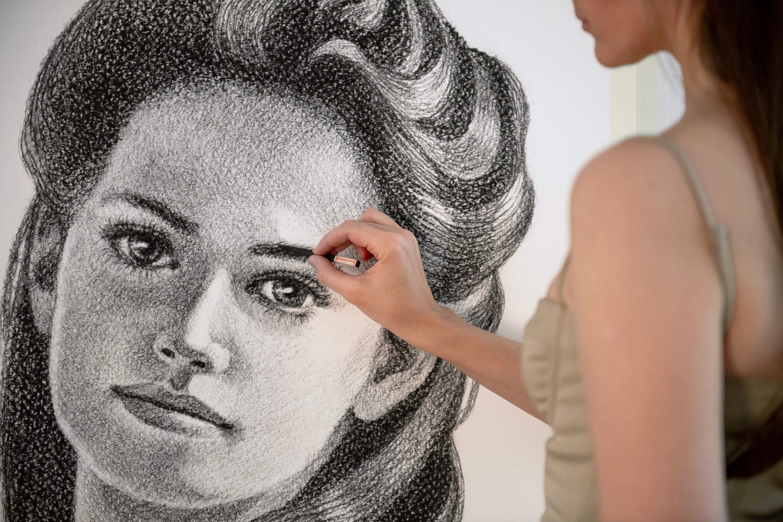 Atlanta Weiss working on a large monochrome portrait drawing of a woman, adding detail around the eye.
